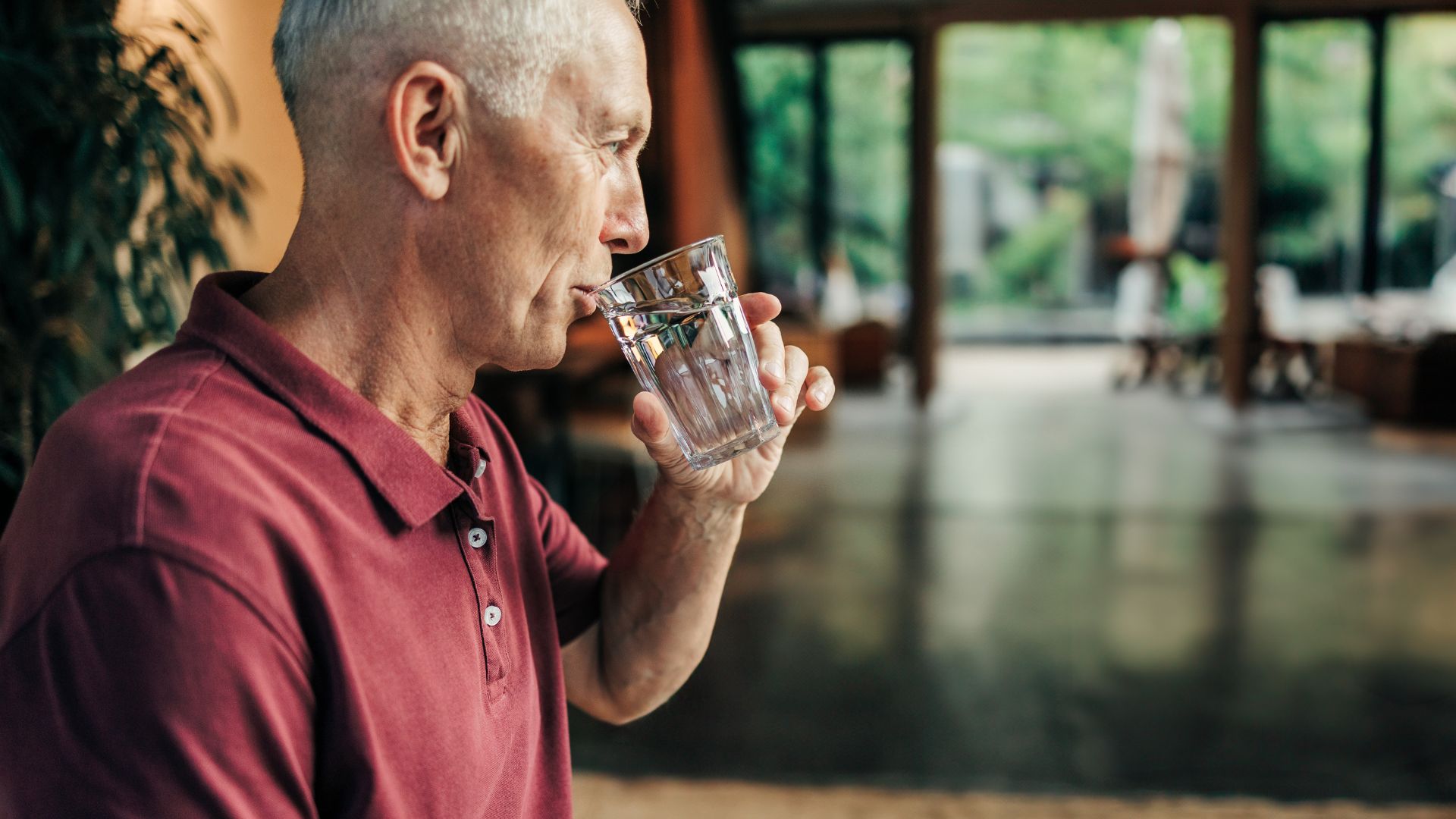 A gray-haired man in a red polo shirt drinks a glass of water during the day to avoid bathroom trips at night.