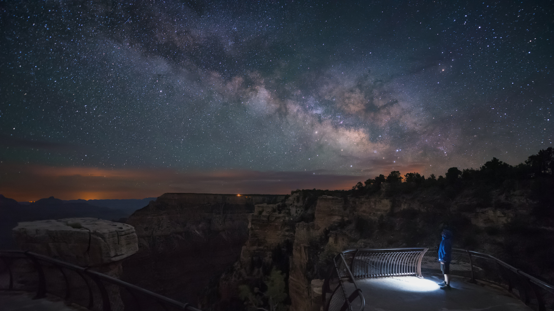 A person wearing a blue coat holding a flashlight stands on a railed balcony overlooking a rocky landscape with the glowing Milky Way in a blue night sky above