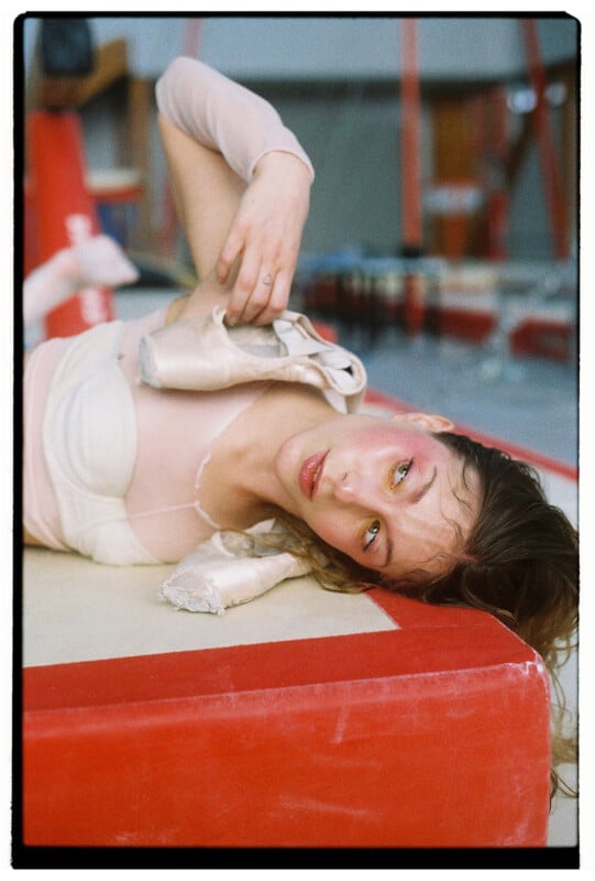 A young woman in a pale leotard lies on a red gymnastics mat, ballet shoes draped over her shoulder. She gazes at the camera with a tired, contemplative expression, and there are gymnastic equipment in the blurred background.