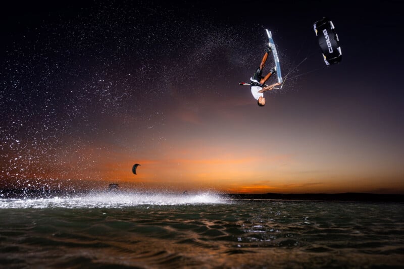 A person kiteboarding performs an aerial trick above the water at sunset, with water spray and another kiteboarder visible in the background.