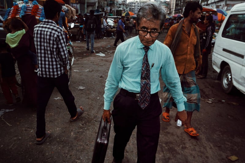 An older man in a turquoise shirt and tie walks purposefully through a crowded, busy street, holding a briefcase. People, vehicles, and market stalls surround him among city chaos.