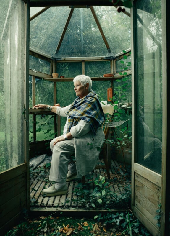 An elderly woman with short white hair sits on a wooden chair inside a small, glass greenhouse, surrounded by plants. She wears light clothing and a blue-green plaid shawl, looking outside through an open door.
