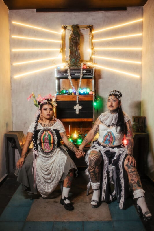 Two tattooed people in elaborate, Virgen de Guadalupe-inspired outfits sit holding hands in a dimly lit room, with a statue and glowing lights arranged behind them, creating a radiant effect.