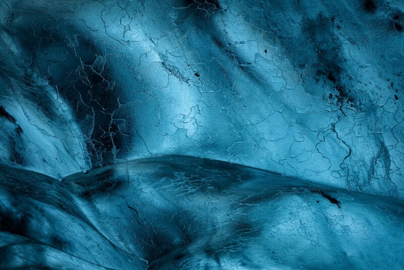 Close-up of a blue, icy surface with smooth curves and visible cracks, resembling abstract frozen terrain or glacier ice with subtle light and shadow variations.