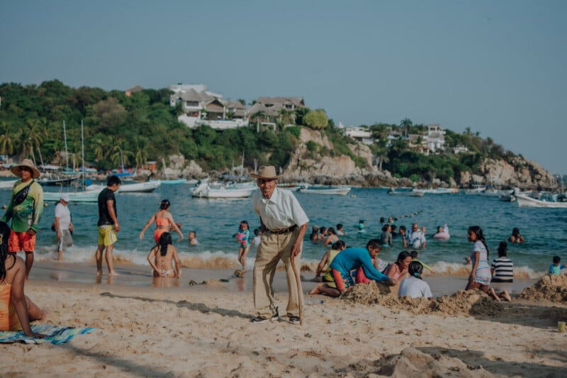 An older man in a hat and dress clothes walks on a busy beach, surrounded by people swimming, building sandcastles, and relaxing. Boats are anchored offshore, with houses on a green hillside in the background.