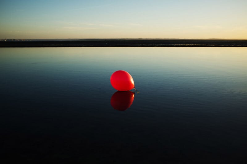A single red balloon floats on calm water at sunset, casting a clear reflection. The horizon and sky are softly lit with fading daylight.