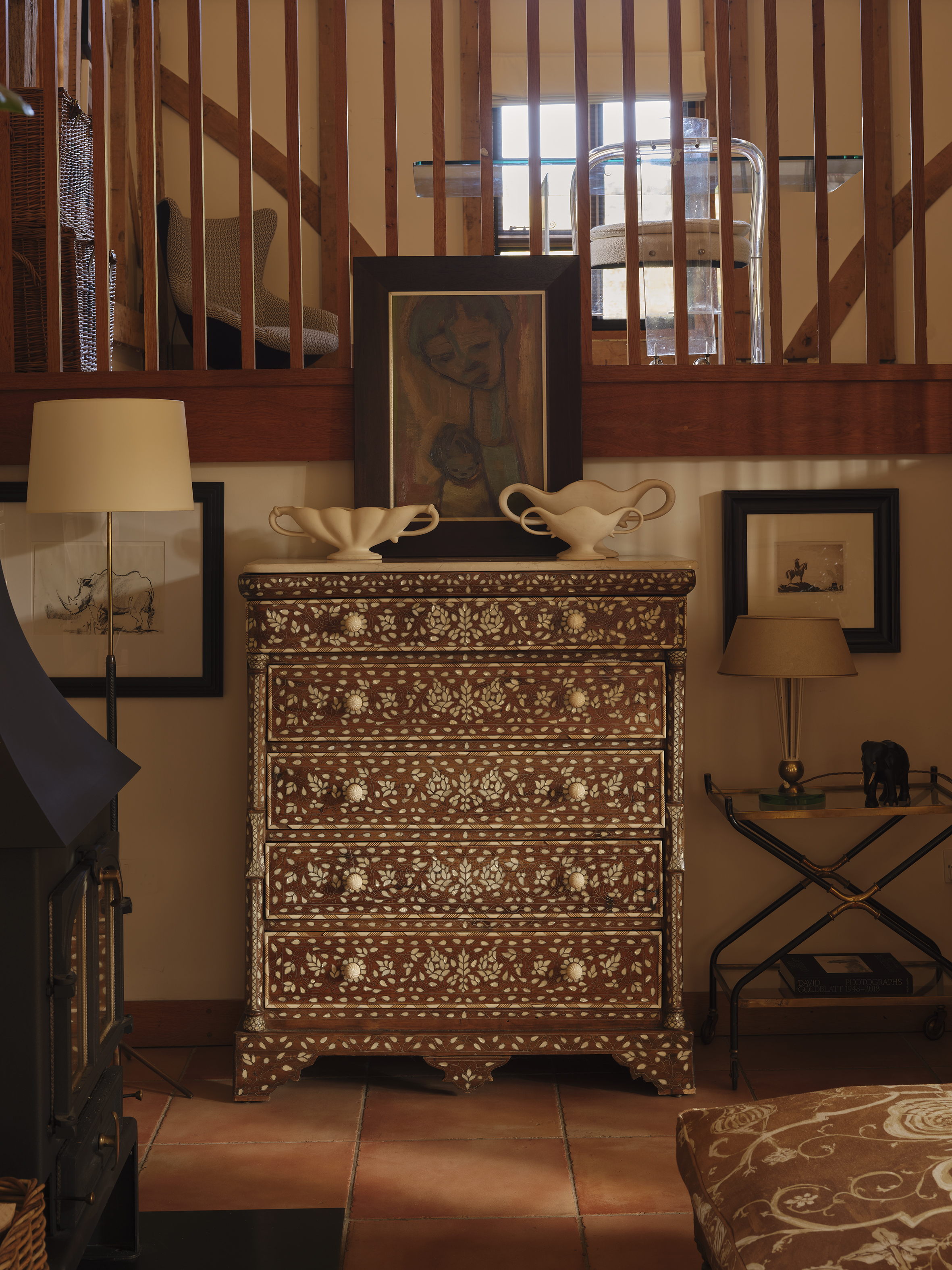 A wooden chest of drawers with intricate floral inlay patterns, topped with a framed painting and two ceramic vessels, in a room with terracotta tile flooring and a wooden balustrade.