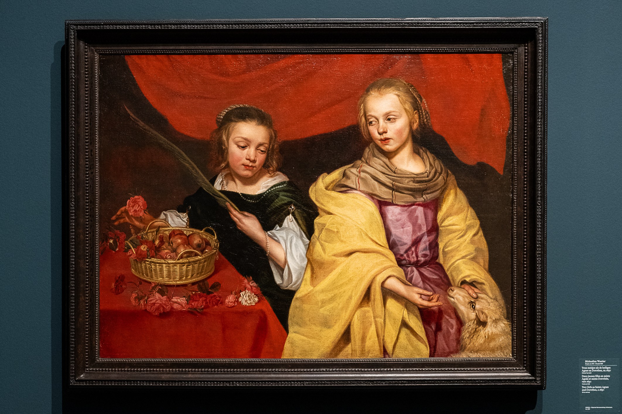 Painting titled "Two Girls as Saint Agnes and Saint Dorothy" by Michaelina Wautier, featuring two young women with symbolic objects, displayed on a dark blue wall in a museum.