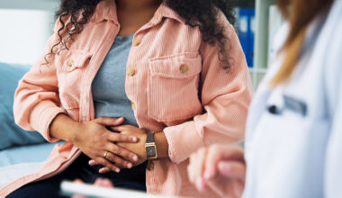 A stock image of a woman seated with her hands across her stomach, while speaking to a medical professional