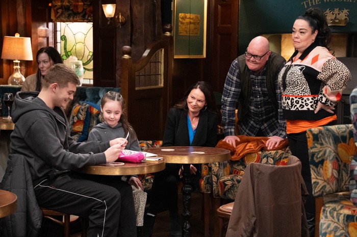 02_04_emm_dylan_paddy_01 Paddy and Mandy stand over Chas, Eve and Dylan, who is looking at a children's drawing, at a table in The Woolpack pub in a scene from Emmerdale
