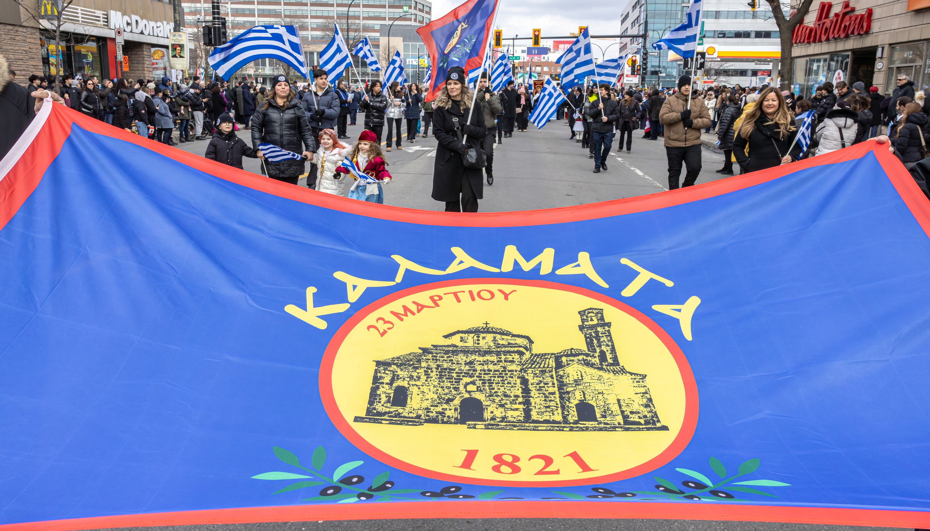 A wide shot of participants in Montreal's Greek Independence Day parade, with a banner in the foreground.
