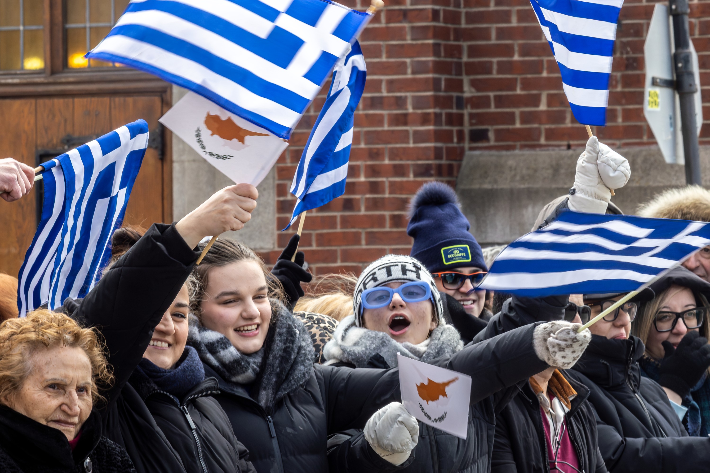 A crowd of parade watchers in winter coats and waving Greek flags.