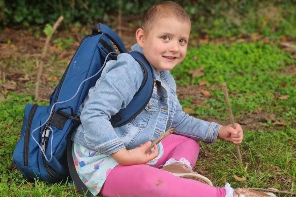 Lacie, 4, is sitting on the grass and smiling at the camera.
She is wearing pink leggings and a denim jacket and holding a stick in each hand. 
She has a large blue medical backpack on with a pipe going into a port. 