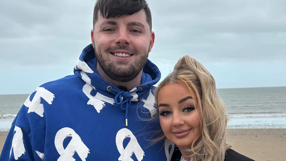 Ruben and partner Amber smiling at the camera on Abersoch beach with the sea behind them. Amber has a long blond ponytail. Ruben is wearing a blue hoodie