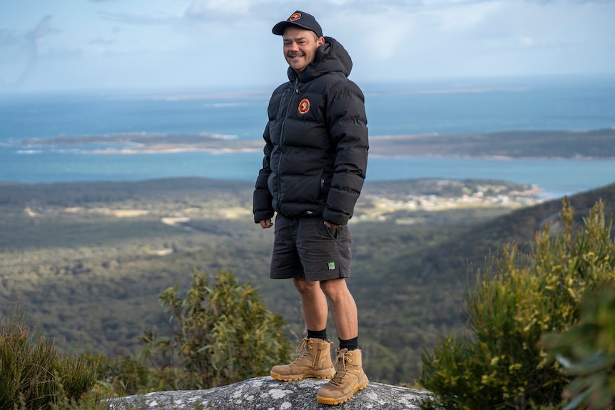 A man in a black hoody stands on a spotty rock with the ocean and a distant island village in the background.
