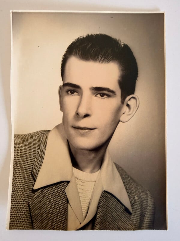 A vintage black-and-white portrait of a young man with short dark hair, wearing a patterned jacket, a wide-collared shirt, and a textured sweater, looking slightly to the side against a plain background.