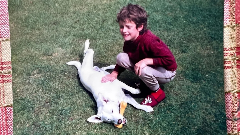 A grainy film photograph taken in the 1980s of a young boy playing on the grass with a white dog, who has a rubber bone in its mouth. The boy has short curly dirty blond hair and is wearing a red jumper, red slippers, and grey jogging bottoms. His eyes are closed and he is smiling.