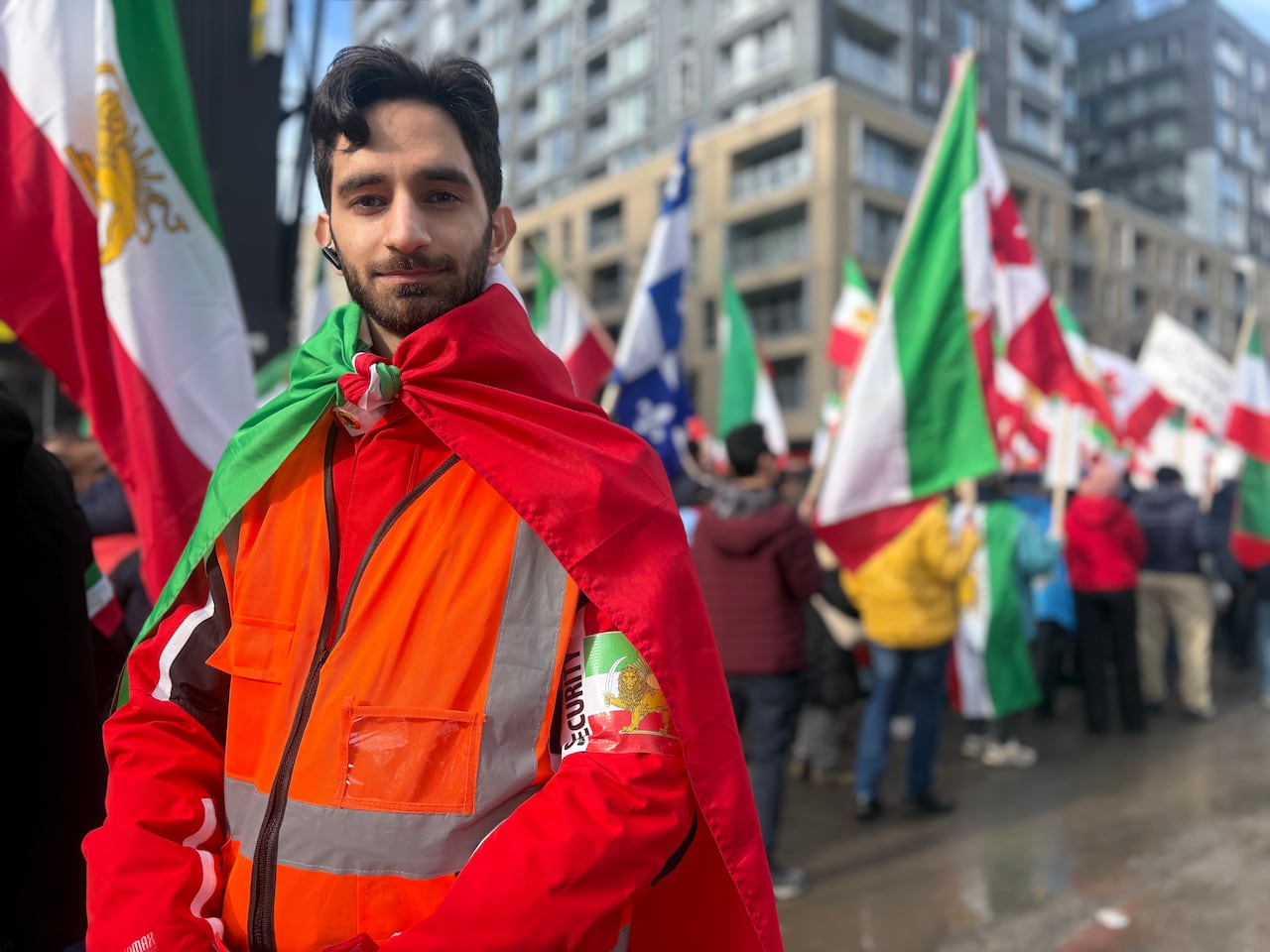 A man attending a rally in Montreal faces the camera and has a flag draped on his back and knotted at the neck.