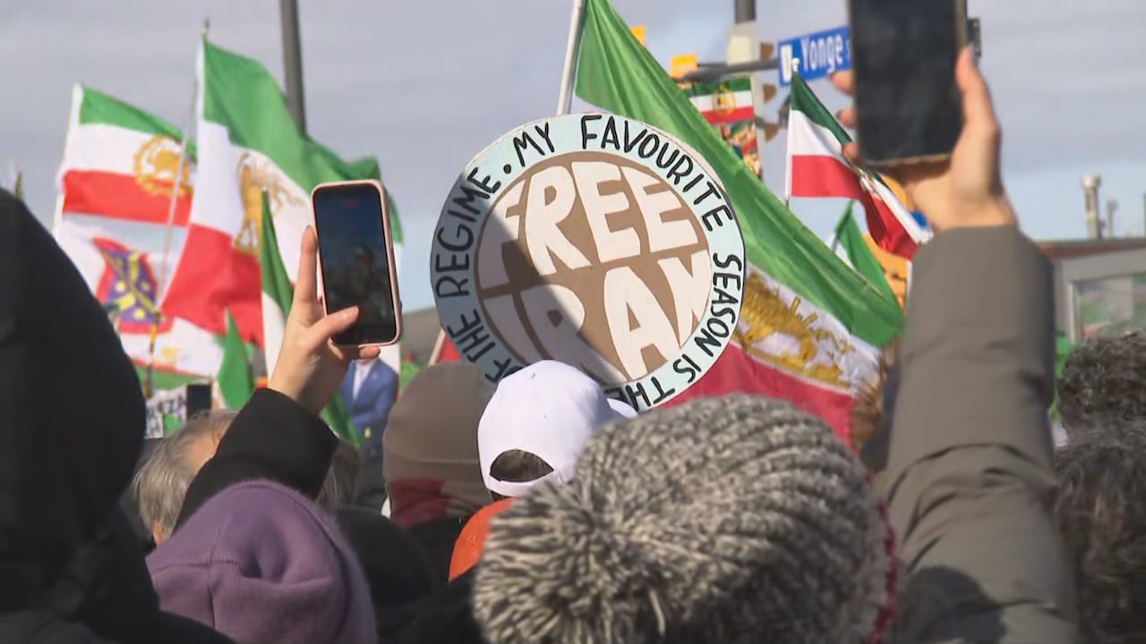 Person holding "Free Iran" sign in crowd with other flags and signs, people holding up phones to film or photograph