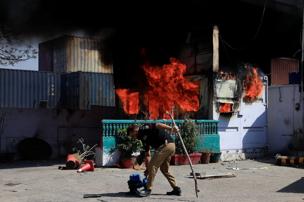 Fire burns in the background as a police in uniform bends over to pick something up from the ground.