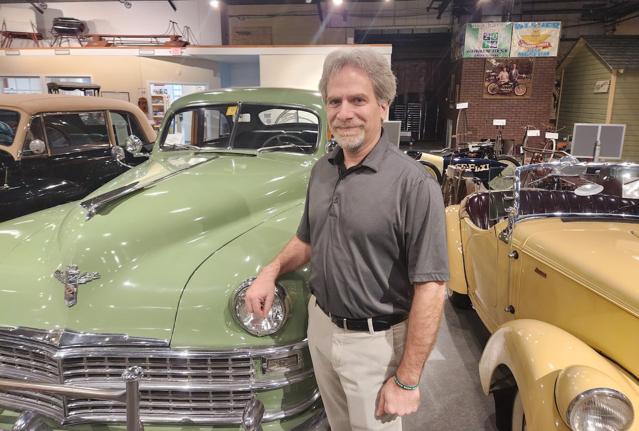 A man with a moustache and goatee poses next to an antique car.