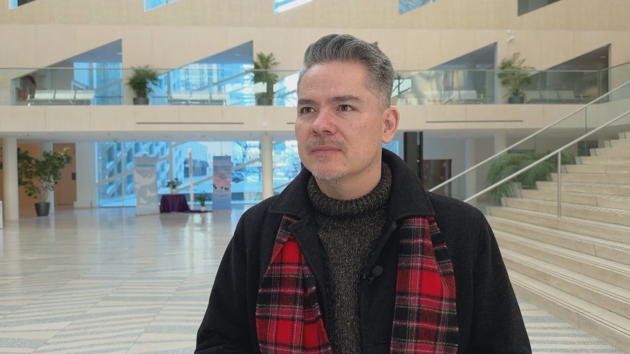 A man with grey hair wears a dark grey sweater, black peacoat and a red plaid scarf. He stands in an empty foyer of the Edmonton city hall building.
