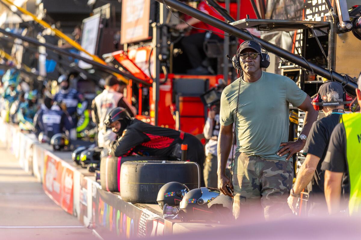 23XI Racing co-owner Michael Jordan wears a ballcap and a headset while standing in the pit at the edge of a racetrack