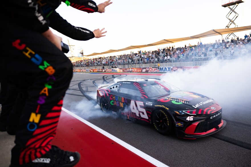 On the far left people are shown from the neck down holding up three fingers as a NASCAR racing car is shown among smoke at right at a race track. 