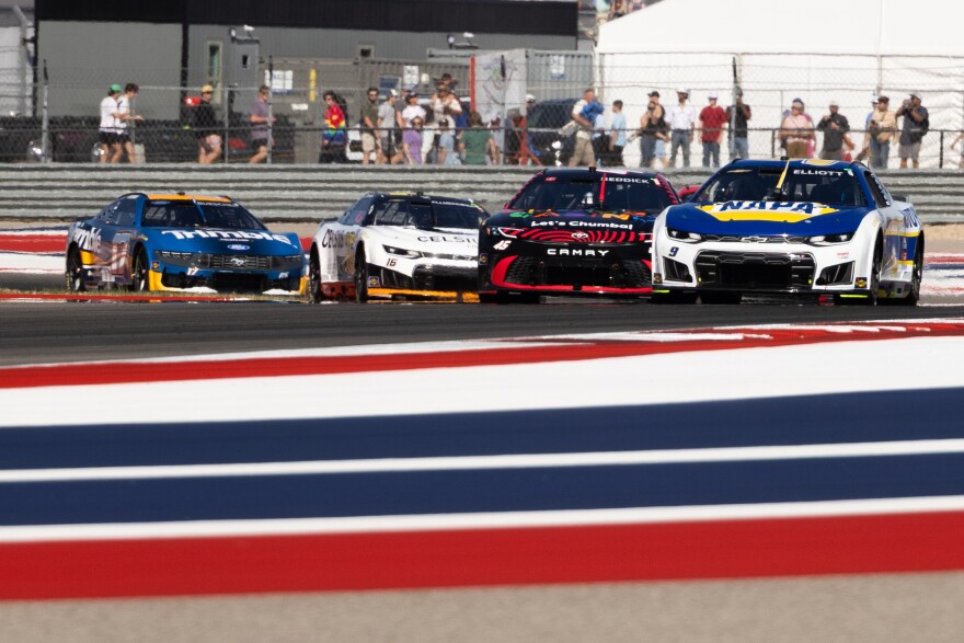 NASCAR cars race around a corner at a racetrack that has red, white and blue stripes in the foreground. 
