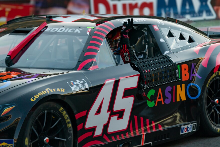 A NASCAR racer holds up three fingers from the window of his car. 