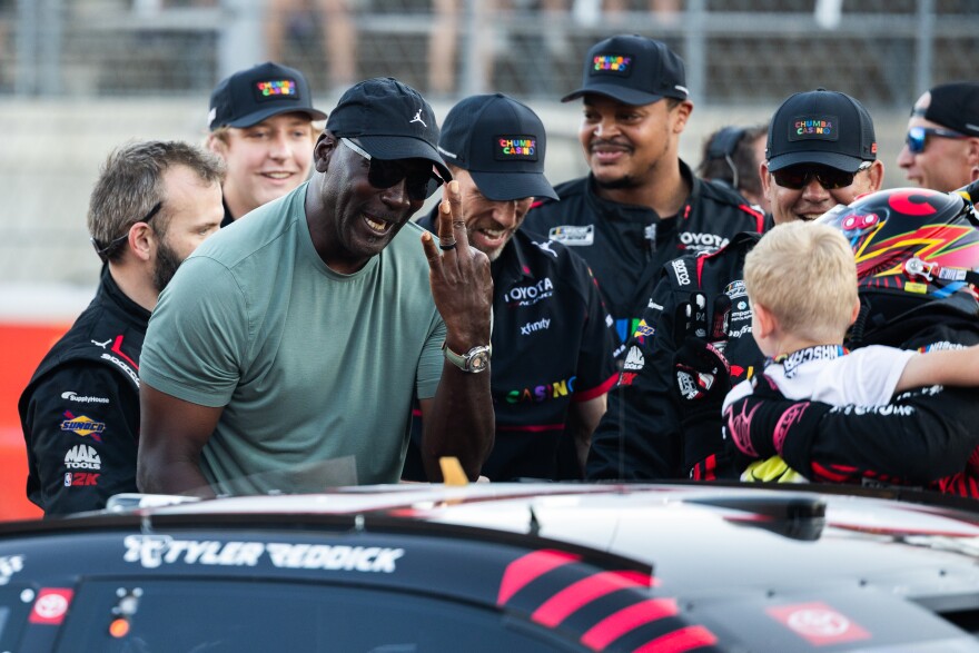 A man holds up three fingers and looks at. a race car driver who is holding a young child during a celebration with other team members. 