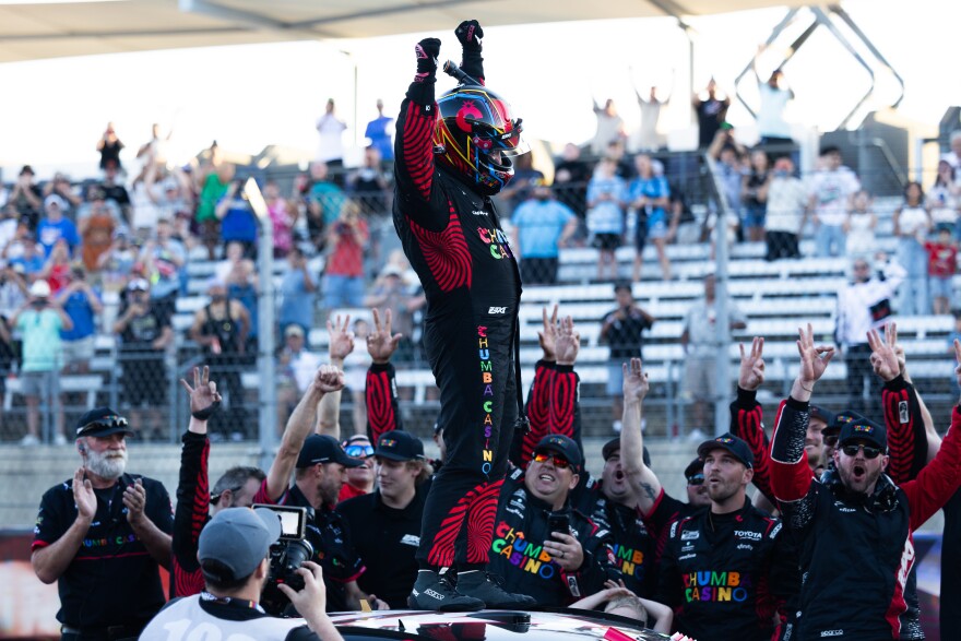 A man in a helmet and racing suit stands on top of a car in front of a big crowd with his arms raised in the air. 