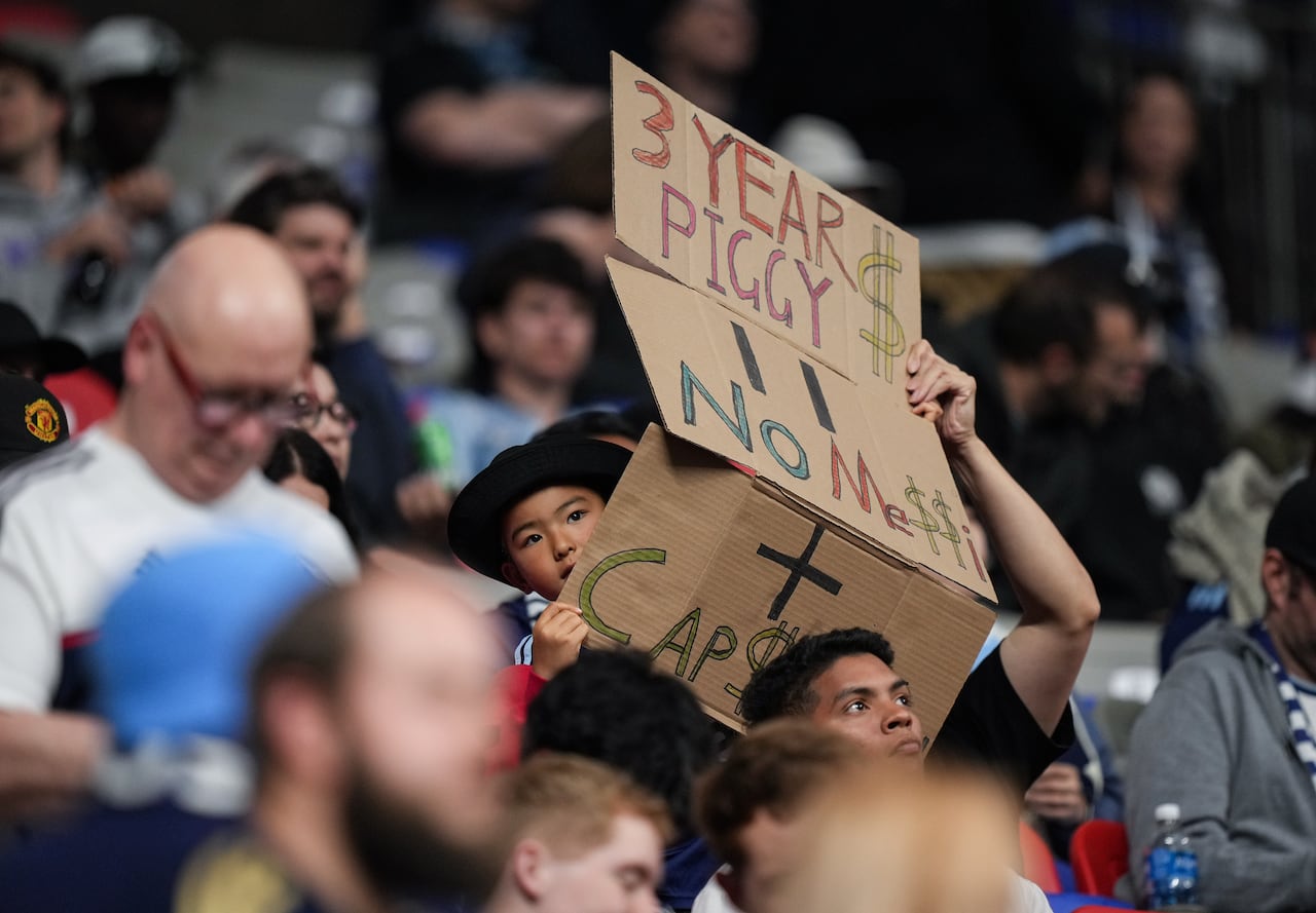 A young fan holds up a sign in a crowd reading '3 year Piggy $' and 'No Messi'.