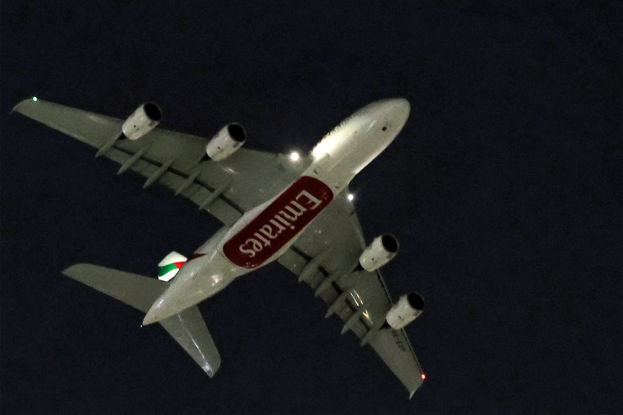 A photo looking up to the underside of a large passenger jet, with the word "Emirates" on the belly, flying in the dark night sky.