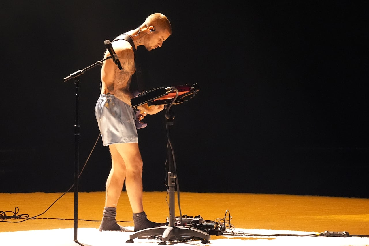 A shirtless man in shiny shorts and socks plays guitar on a stage. 