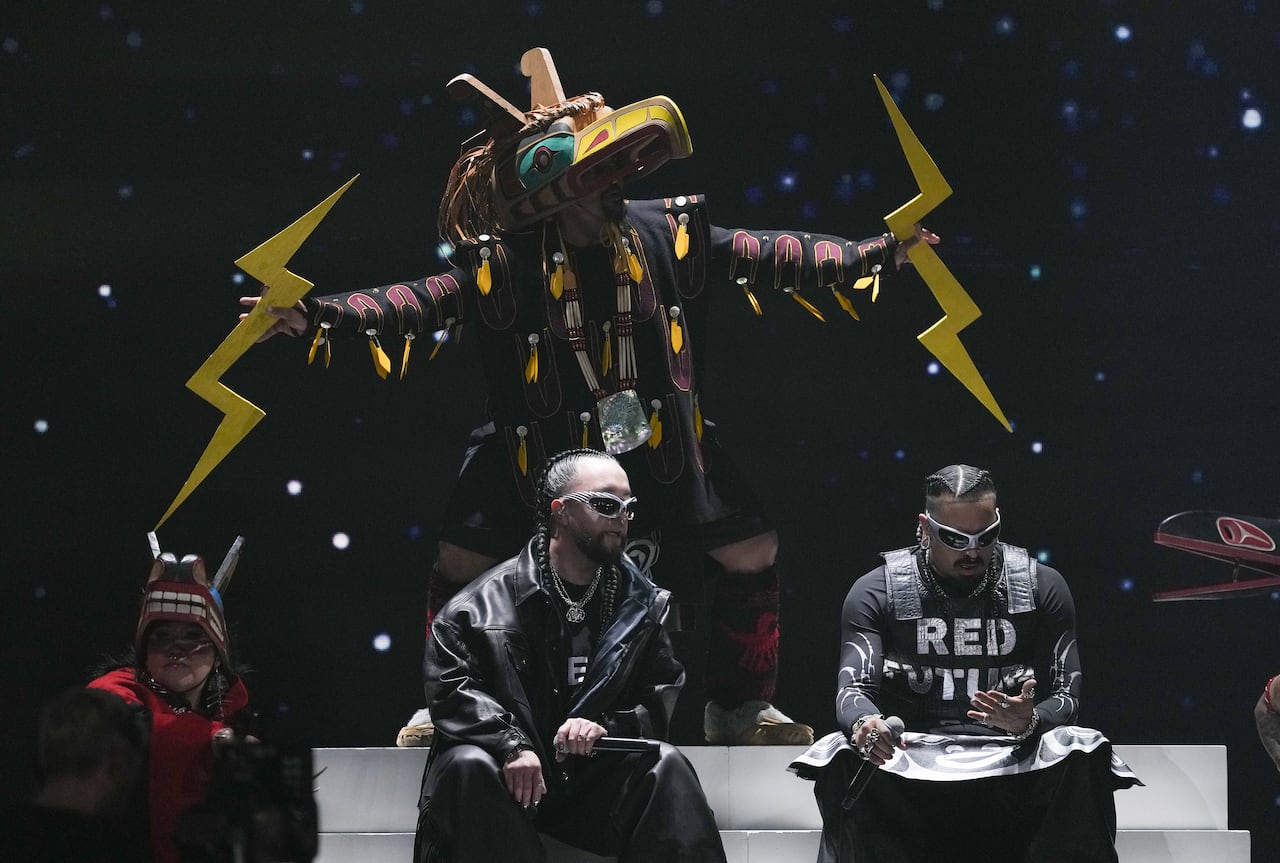 Two people sit on a stage while someone in a costume inspired by Indigenous cultural dress dances behind them.