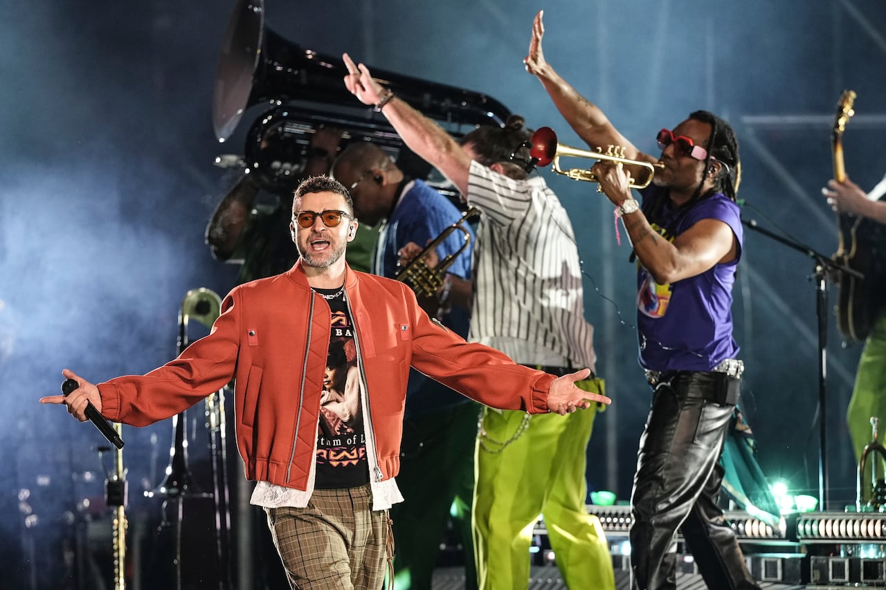 A man in a red jacket and brown plaid pants, wearing sunglasses, stands on a concert stage with his arms outstretched and a microphone in his right hand as he sings, while a band playing instruments marches behind him.