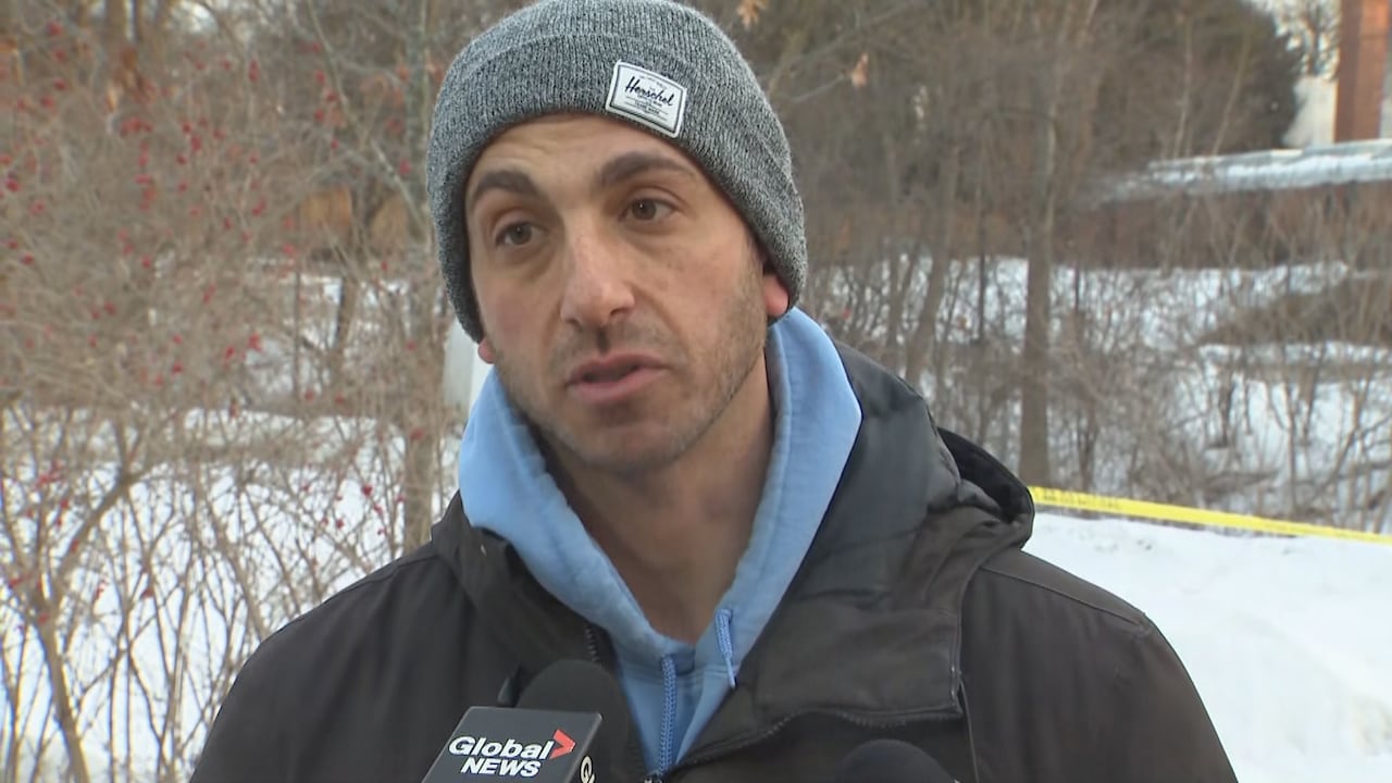 A middle-aged man in a grey toque, blue hoodie and black winter jacket speaks into a Global news microphone in front of a taped off synagogue in winter