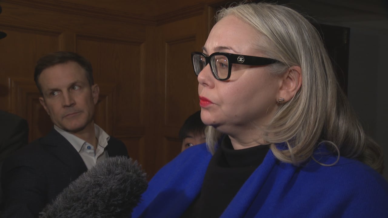A white woman with white hair speaks to a gaggle of mics, with a white man next to her.