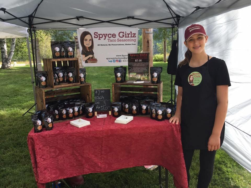 A smiling girl next to a farmer's market stand showcasing packets branded "Spyce Girlz"