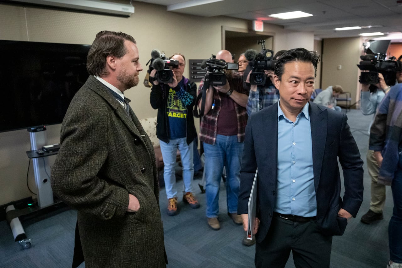 A man wearing an overcoat looks on as another man wearing a suit turns away from him, with a gaggle of camerapeople visible.