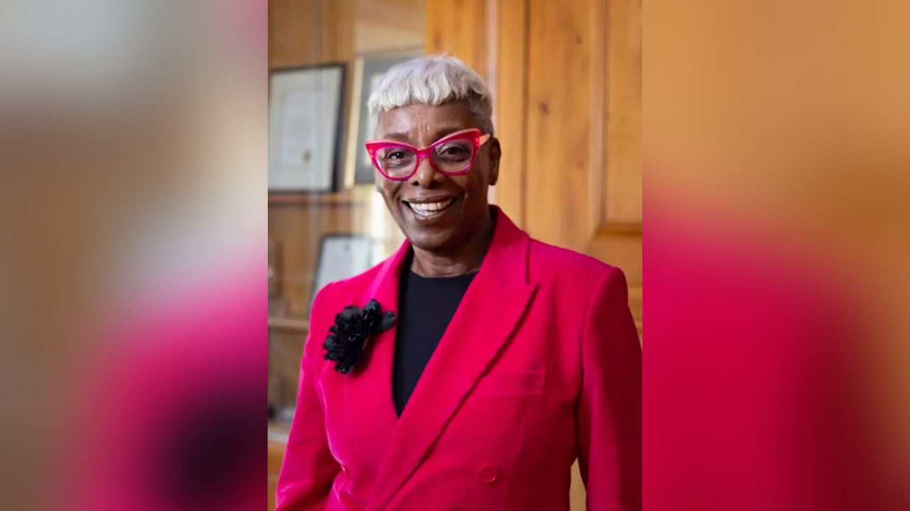 A Black woman with short white hair, wearing glasses with bright red frames and a hot pink blazer over a black top, smile as she poses for a portrait in an office with light brown wood panel walls in the background.