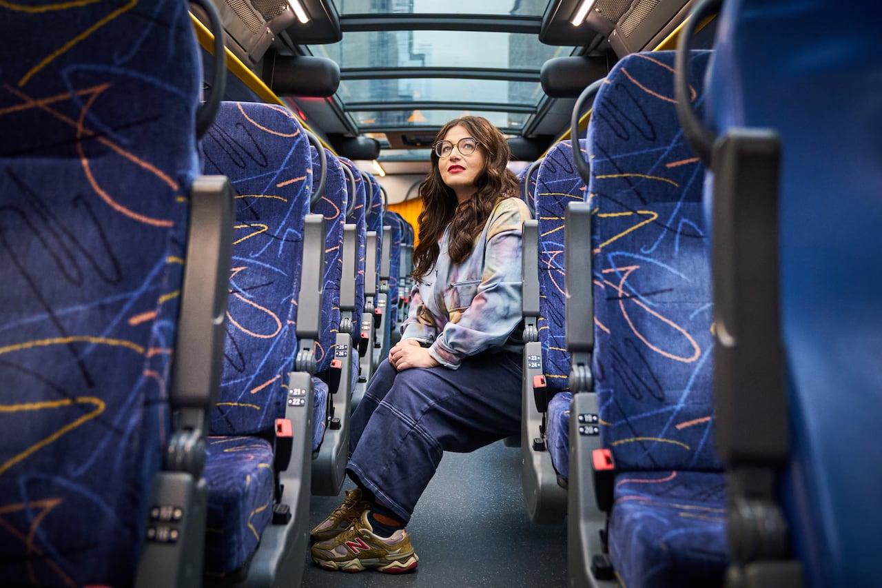 A woman with long brown hair and glasses, wearing a light blue denim jacket with a grey hood and blue jeans, poses for a photo sitting inside a bus with blue seats and blue carpet.