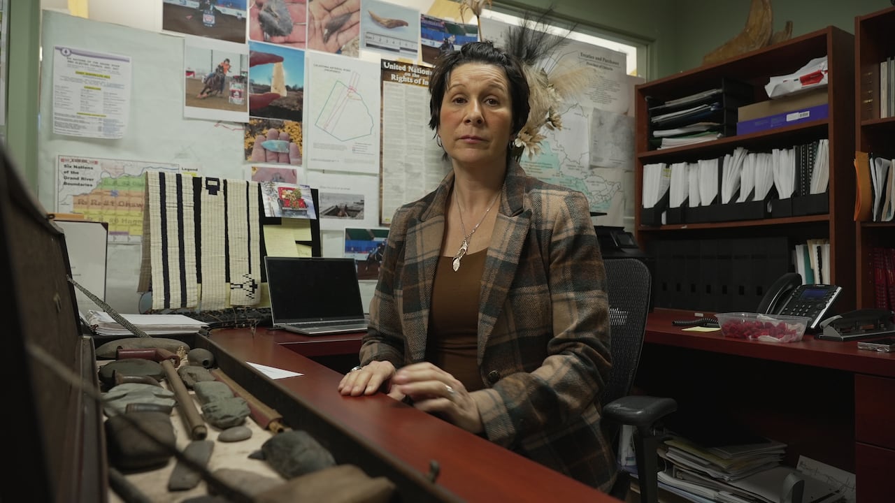 An Indigenous woman with very short, dark hair sits in front of an office desk with an open rectangular box holding Indigenous artifacts in front of her.