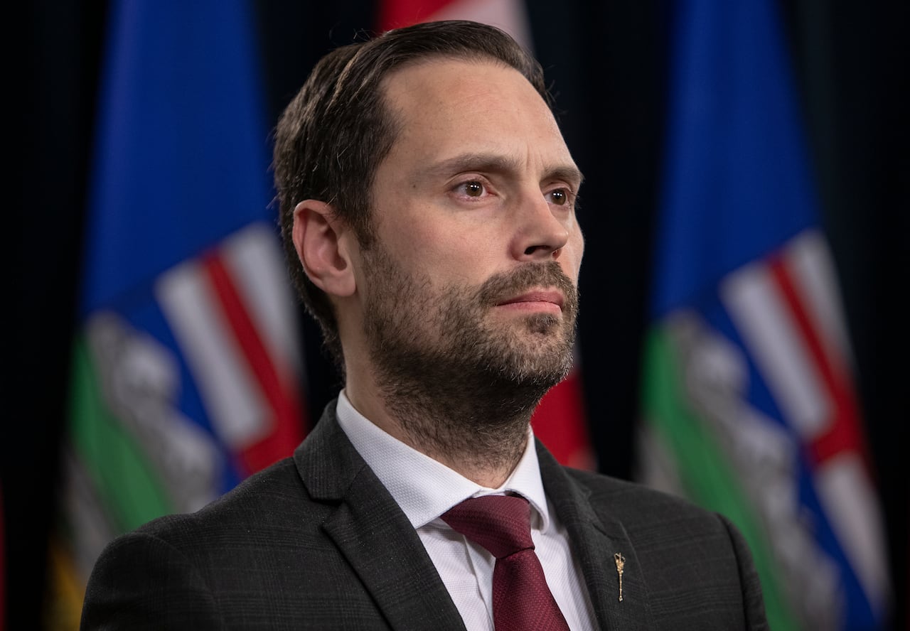 A man with a beard and wearing a black suit and burgundy tie looks off-camera. Alberta flags are in the background.