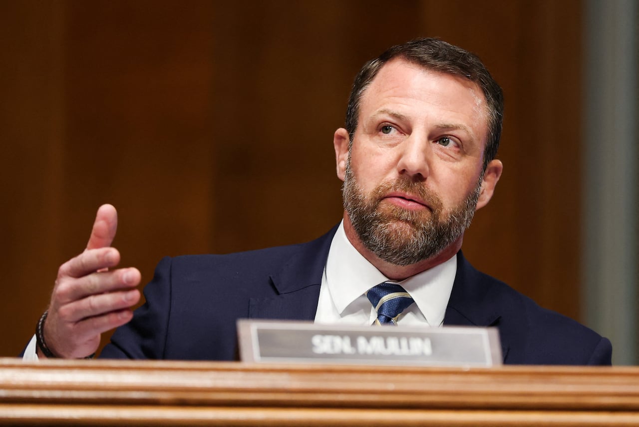 Man in blue suit sits at a desk with name in front of him 