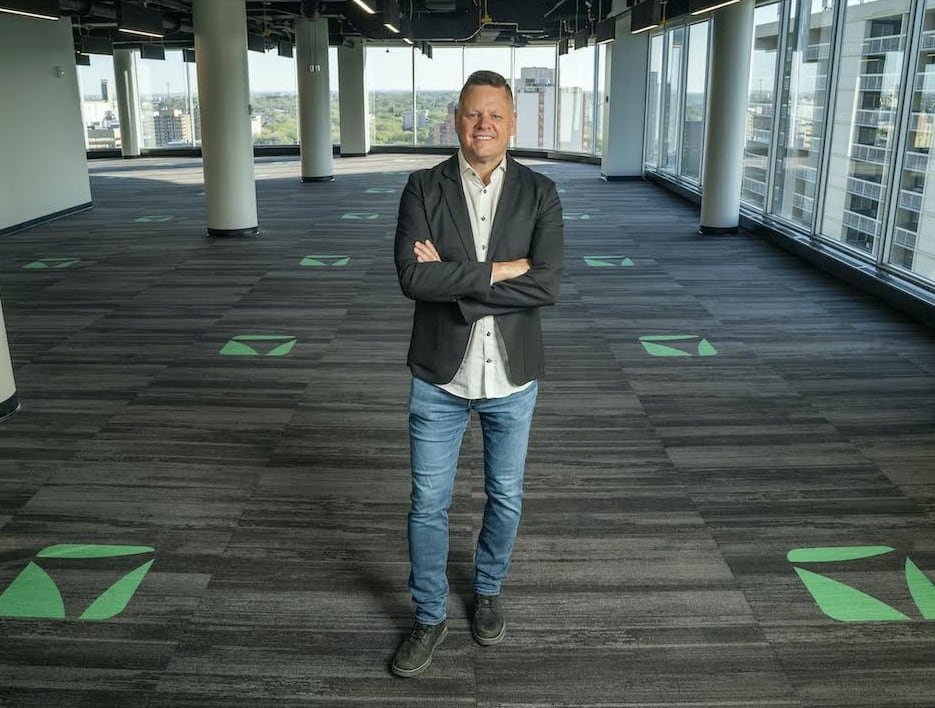 A man in a sport coat and jeans stands in an empty office open-plan office with a green logo on the carpet.