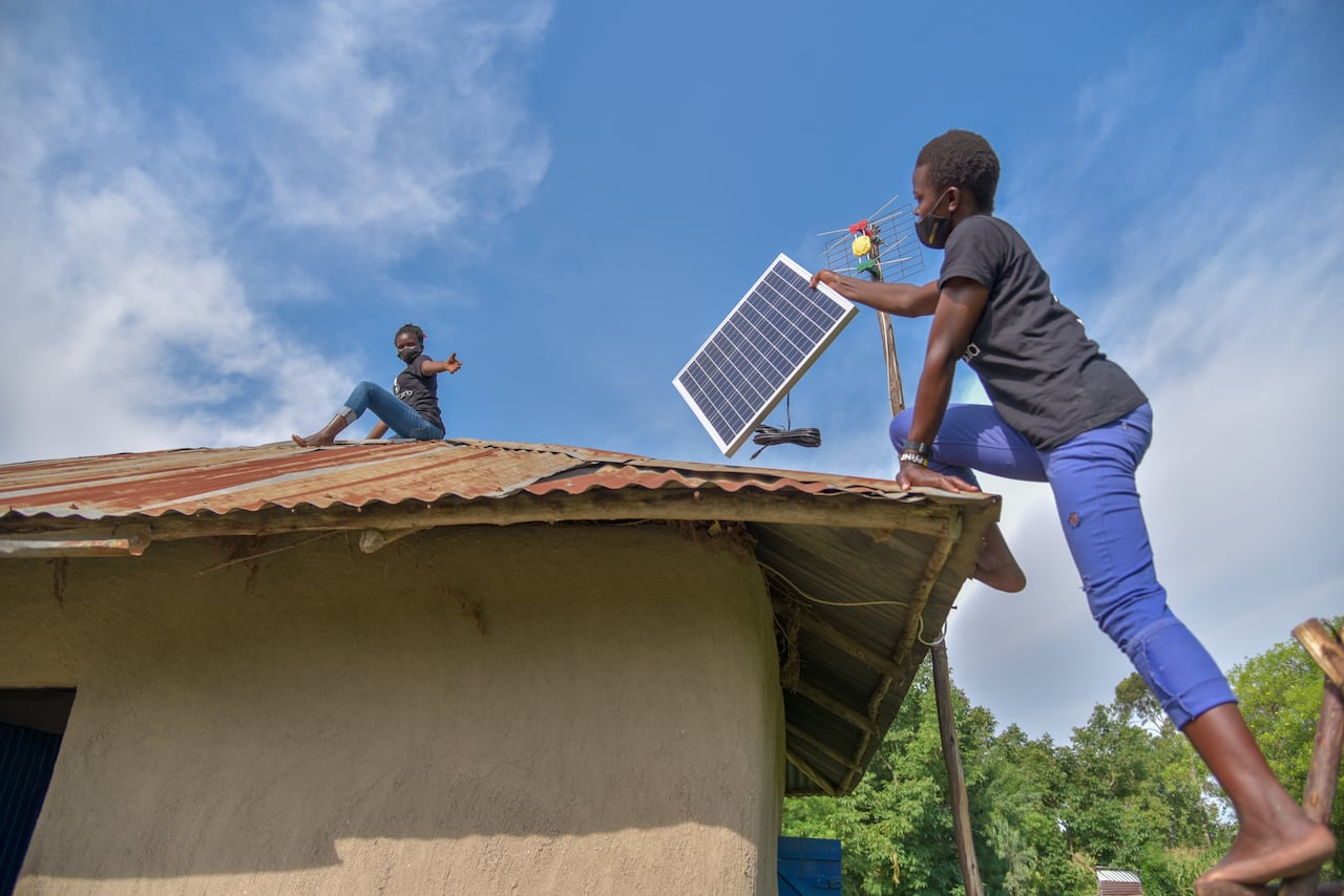 Woman hands a small solar panel to a woman sitting on a roof