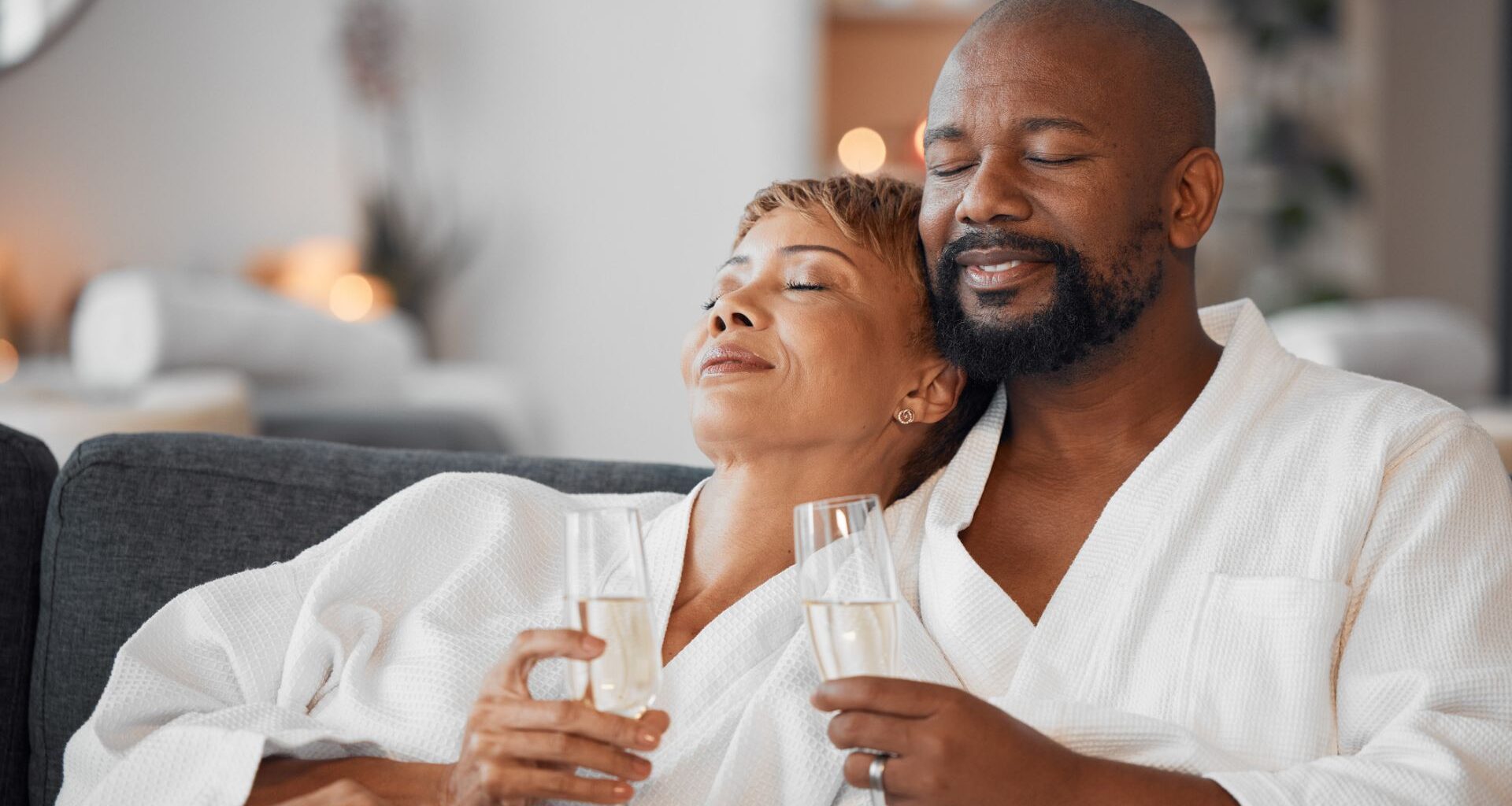Wealthy middle class couple in robes, relaxing on a spa couch with champagne.