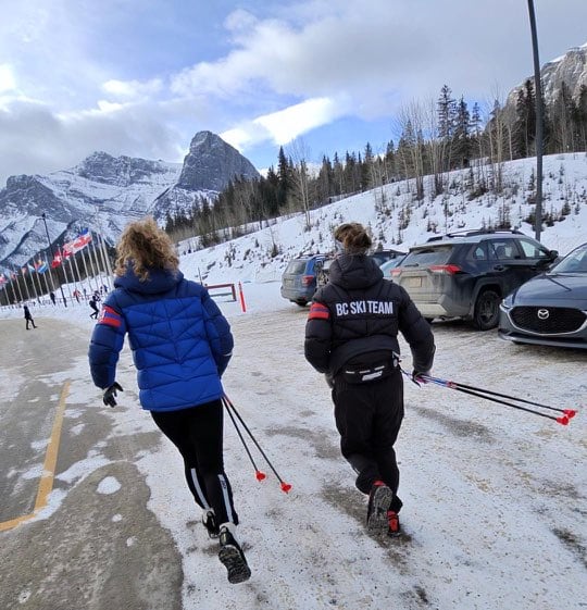 Two girls run together holding ski poles with mountains in the background